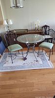 Full view of round glass top kitchen table with two metal framed chairs with upholstered seats placed on a rug.