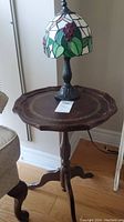 Photo showing slag glass lamp with grape and leaf design shade on wooden pedestal table with carved tripod legs.