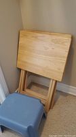 Two wooden TV trays folded and leaning against the wall, with a small blue plastic stool in front on carpet.
