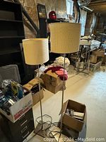 Two IKEA floor lamps with white drum shades and brushed metal stands in a basement storage area with other assorted items around.