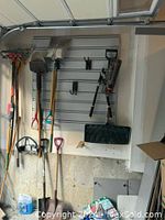 Garage wall with various gardening tools including rakes, shovels, and garden hoe hung on a panel, with some gardening supplies on the floor.