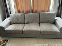 Photo of a grey Urban Barn modern sofa with three seat cushions and three back cushions, placed in front of windows with curtains.