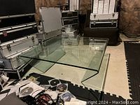 Full view of modern glass coffee table showing the square top and bent glass legs, placed on a light floor with cases in background.