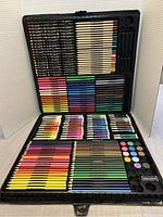 Opened deluxe art supply case showing various compartments filled with colored pencils, markers, oil pastels, crayons, watercolor paints, eraser, and sharpener.