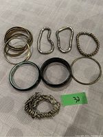 Bracelets arranged on beige fabric showing various types and styles including gold-tone bangles, silver-tone linked bracelets, black and blue solid bangles, silver-tone beaded bracelet and chunky chain link bracelet.