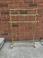 Front view of the brass plated blanket rack showing the two horizontal bars and height relative to ground and brick wall background.