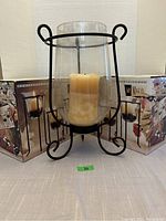 Large black metal candle holder with glass cylinder and beige pillar candle inside, shown on table with box packaging in the background.