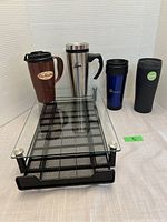 Set of four assorted coffee travel mugs with a metal mesh coffee pod drawer beneath them, displayed on a tabletop.