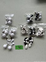 Photo showing 10 pairs of earrings grouped in small sealed plastic bags, arranged on a grey surface. Two main designs visible: predominantly white bead design and mixed black and white bead design.