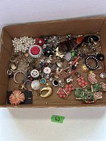 Box of assorted costume jewelry pieces including brooches, bracelets, earrings, chains, and pins.