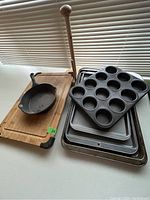 Photo showing cast iron skillet on wooden cutting board alongside metal baking sheets and muffin tin stacked on counter in natural light.