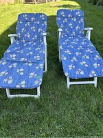 Two white plastic lounge patio chairs with blue floral cushioned pads positioned on grass, showing overall front view and reclining position.