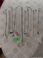 Photo showing 14 silver-tone necklaces laid out, each with different charms including wings, butterflies, cross, floral, and letters.