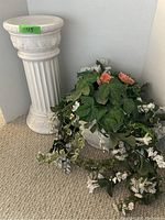 Photo showing white tall pedestal with fluted design and white flower pot filled with artificial flowers and greenery on carpet.