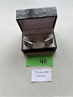 Full view of stainless steel cuff links inside the black textured box with pink lining, displayed on a light surface.