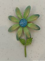 Close-up of the tin flower brooch showing the blue textured center, green petals with blue shading, and two green leaves on the stem.