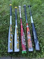Five used youth aluminum baseball bats lying on grass showing overall condition and details including brands and wear.