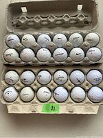 Top-down view of 24 used golf balls arranged in three flat egg cartons showing variety of brands and visible signs of wear and dirt marks.