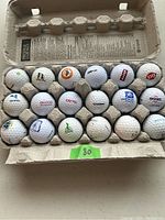 Photo showing all 18 golf balls in a cardboard carton with visible printed logos from multiple companies