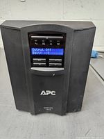 Front view of APC Smart-UPS 1500 UPS showing LCD screen with status message 'Output Off Batt: --%' and control buttons below.
