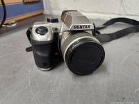 Silver Pentax digital camera with lens and strap, shown from front-left angle.