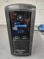 Front view of the CyberPower 1000VA AVR UPS showing power button and LCD display with load, battery capacity, and voltage reading.