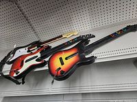 Three guitar controllers on a shelf, showing detailed colors and button layouts of each, including straps and different colored fret buttons.