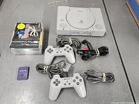 Photo showing PlayStation 1 console, two controllers and cables, stack of game cases, and memory card on a grey surface.