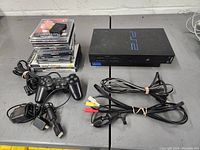Full lot shown including PlayStation 2 console, controller, power cord, AV cables, memory card, and stack of game discs for PS1 and PS2.