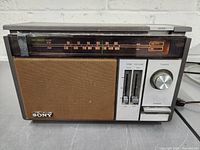 Full view of Sony 2 band AM/FM radio showing speaker, tuning dial with frequency scales, tone and volume sliders, tuning knob, and power switch