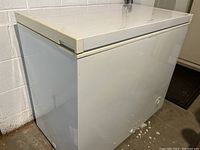 White chest freezer angled to show front and left side, in a garage setting.