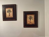 Two framed palm tree prints hanging side by side on a white wall. Both frames are dark brown with heavy distressing and vintage-style palm tree botanical illustrations.