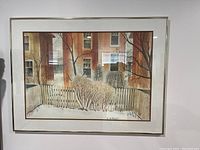 Framed print showing a winter scene with bare bushes, white picket fence and apartment building windows in background. Signed artwork visible in corner. Matte and simple white frame with glass.
