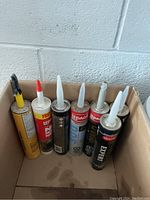 Six tubes of assorted caulking and adhesive in various colors and brands, some with red and black labels, stored upright in a cardboard box.