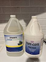 Photo of two commercial cleaning containers, Dynamite Dustbane cleaner and Tuff-Cote Floor Finish, against a brick wall.