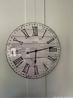 A front-facing image of a large round Westclox wall clock featuring black Roman numerals on a distressed white wooden plank pattern background. The clock shows time around 4:15 and has black hour and minute hands. Brand name visible.