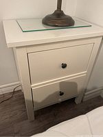 White IKEA end table with two drawers, showing front view with lamp base on top and glass plate.