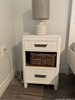 Pair of smaller white painted wood end tables each with two drawers and a middle open shelf with woven basket, showing full front side with lamp on top of one table.