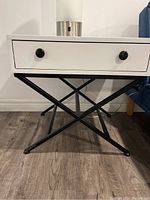 Front view of the modern end table showing the white drawer with black knobs and black steel base with intersecting X design.