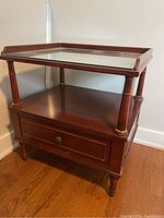 Wooden side table with rectangular glass top, raised sides, single drawer below, turned legs, and dark brown finish.