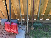 Photo showing multiple garden tools and snow shovels arranged against wooden fence, includes plastic snow shovels and several metal-handled garden implements.