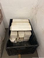 Eight boxes of glass tiles stacked in a plastic crate in a basement corner. Boxes are white and sealed.