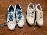 Two pairs of white ladies golf shoes side by side on a wooden floor, one pair with blue shoelaces and blue insoles, the other with white shoelaces with brogue style detailing.