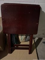 Photo of one folding TV tray with reddish stained wood, folded and standing upright showing surface scratches and wear.