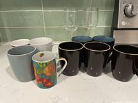 Photo showing various assorted wine glasses and mugs arranged on a kitchen counter including clear wine glasses and multiple mugs of different colors and designs.