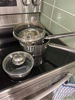 Three stainless steel saucepans stacked on stove, one with a glass lid, one with a strainer insert, with metal handles.