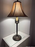 Lamp lit, showing warm light with bronze colored base and brown fabric shade on a glass surface.