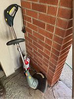 Electric weed trimmer showing full unit leaning against wall with fire extinguisher behind it.