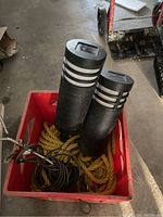 Two black solar path lights, yellow rope, and metal screw-in dog lead coiled in a red container on floor.