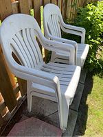 Side angle showing two white plastic chairs with slatted backs and armrests, stacked on a stone patio against a wooden fence and greenery.
